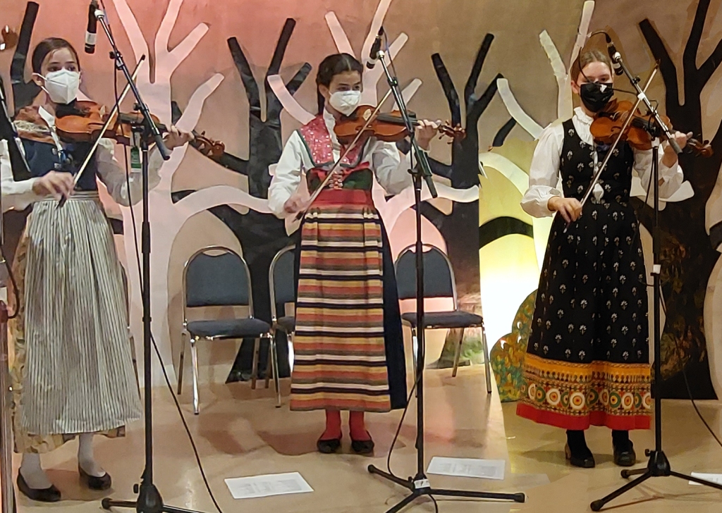 Three youth musicians in Nordic costumes playing violins on a stage decorated with tree outlines. 