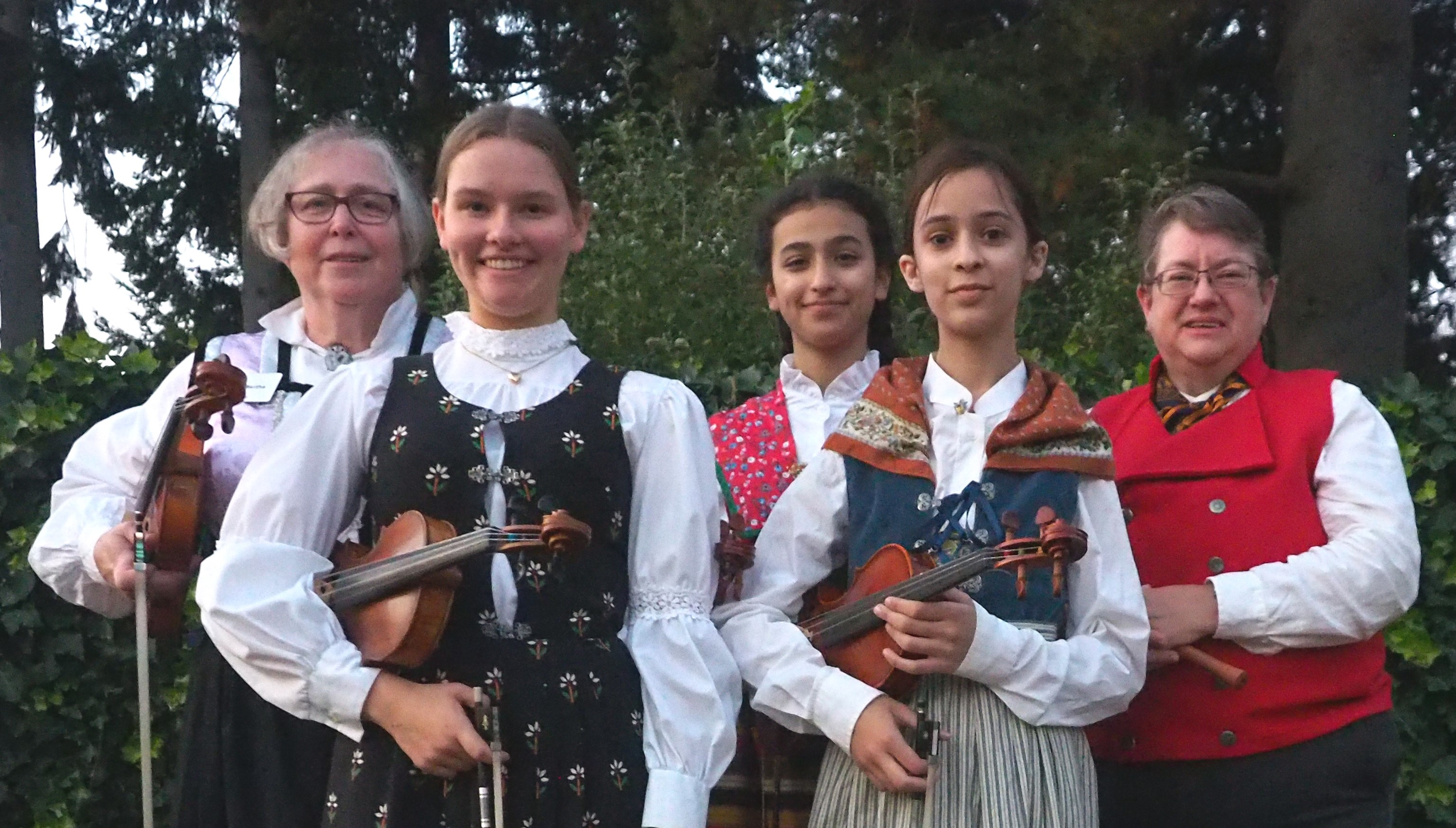 Picture of three youth musicians and two adult musicians, all in Nordic costumes standing in front of trees. 